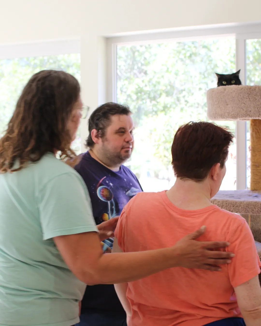 Three people in a sunny room, with a black cat peeking from the top of a cat tree.