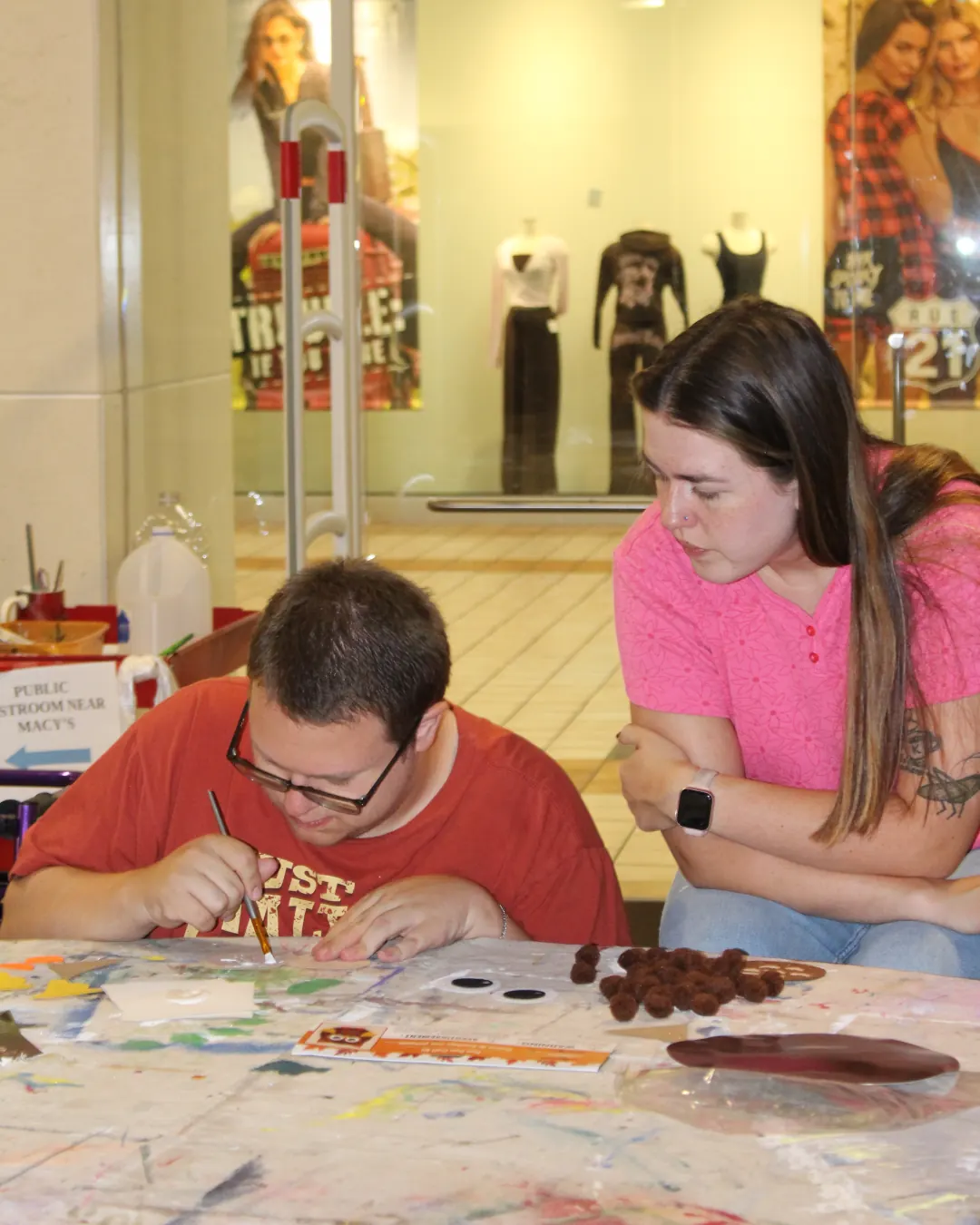Two people at a craft table; one paints a project while the other looks on.