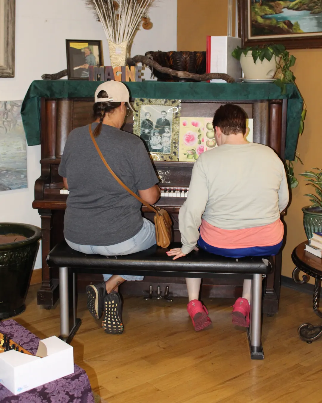 Two people side by side on a bench, playing an old upright piano.