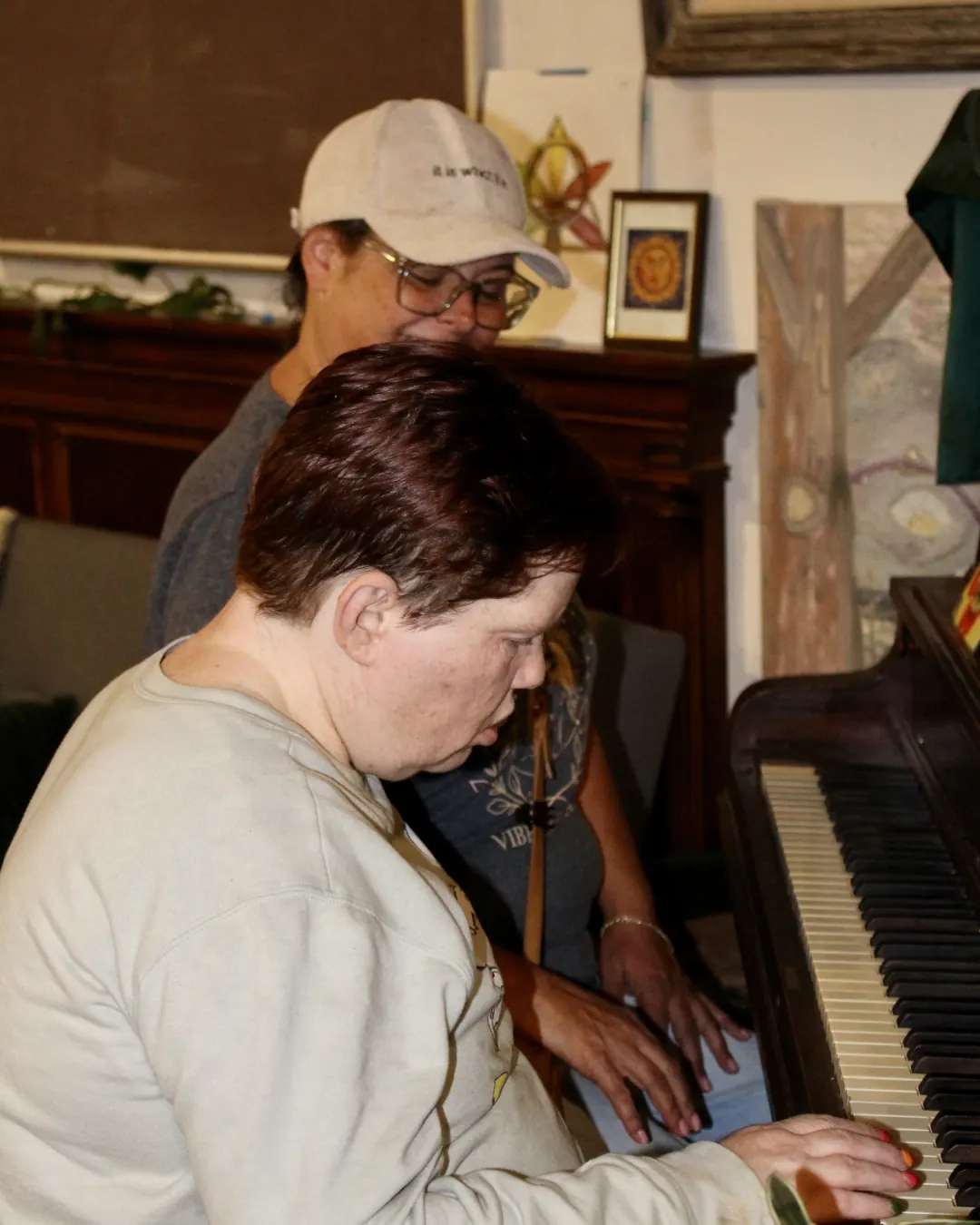 One person plays the piano while another leans in beside them, smiling.